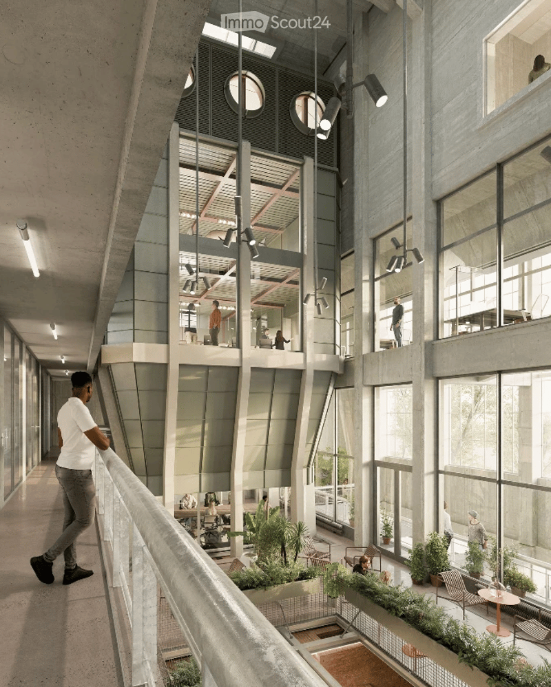 Modern, open-plan corridor with glass walls and a balcony overlooking a lower level. The space features exposed concrete columns, hanging lights, and a few people walking or standing. There are potted plants and seating areas visible in the lower level.