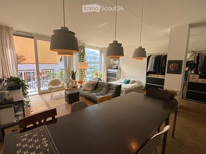 Modern, open-plan living and dining area with large windows and balcony access. Features a sofa, armchair, dining table with chairs, pendant lights, wardrobe, and a bed in the background.