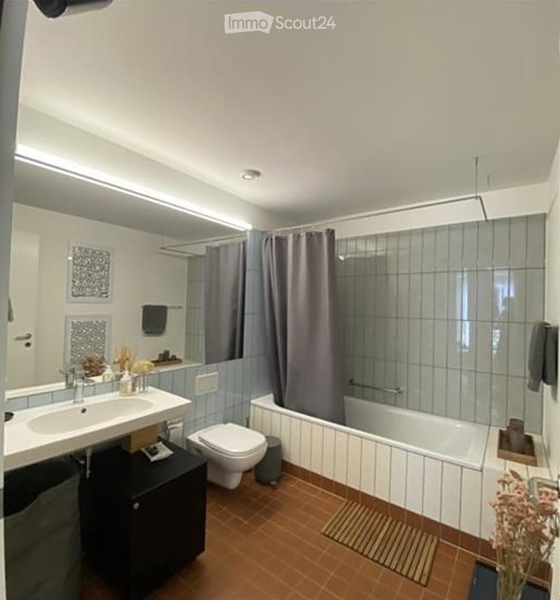 Modern bathroom with a white sink, mirror, toilet, bathtub, and shower curtain. Features a dark wooden cabinet and a small flower vase on the floor.