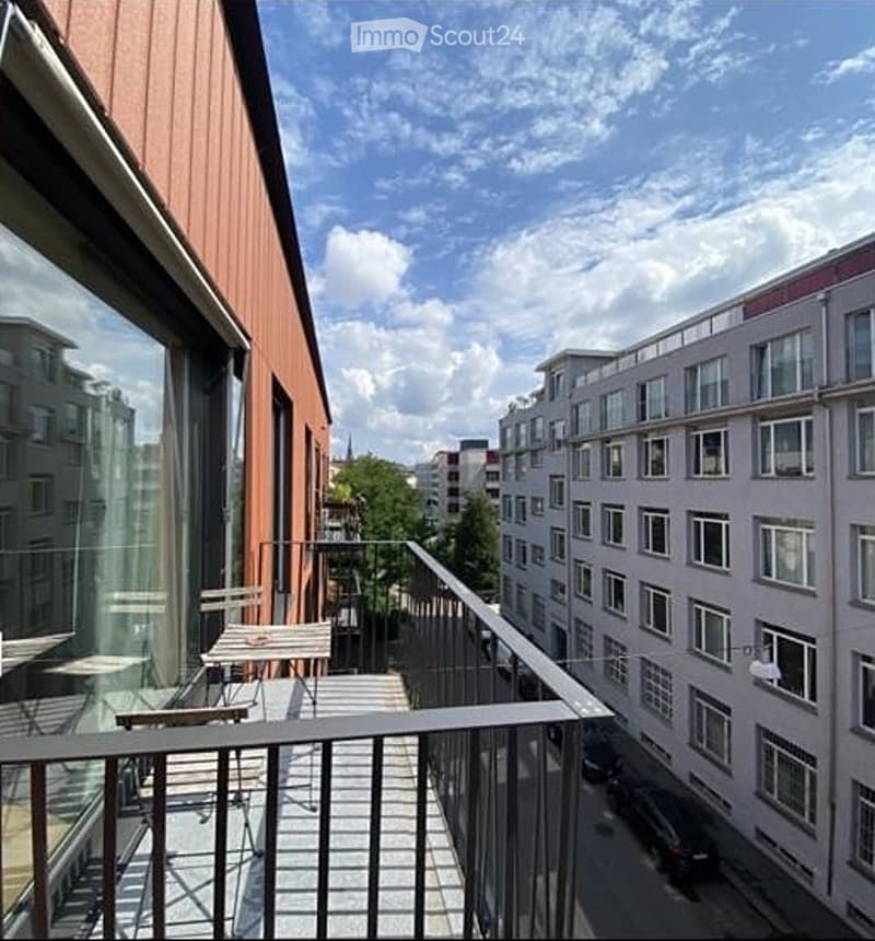 Balcony with metal railing, outdoor furniture including a table and chairs, overlooking a cityscape with modern buildings and trees.