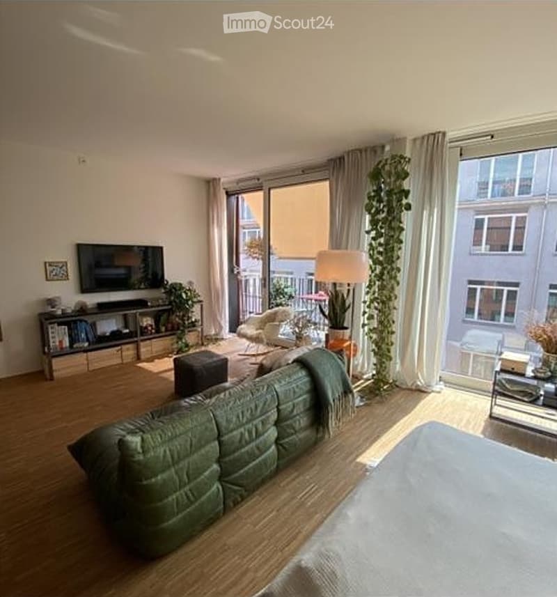 Bright living room with large windows and a balcony, featuring a green sectional sofa, coffee table, armchair, side table, floor lamp, TV, and indoor plants.