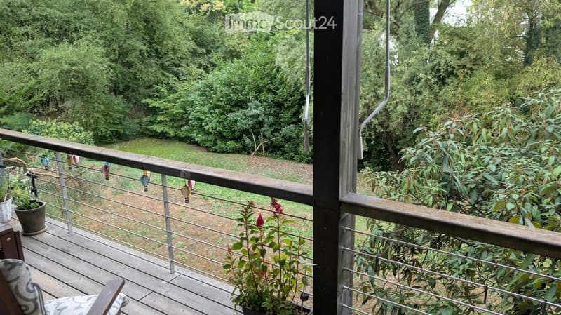 Balcony with wooden decking, metal railing, potted plants, and colorful hanging ornaments. Offers a view of a lush green garden.