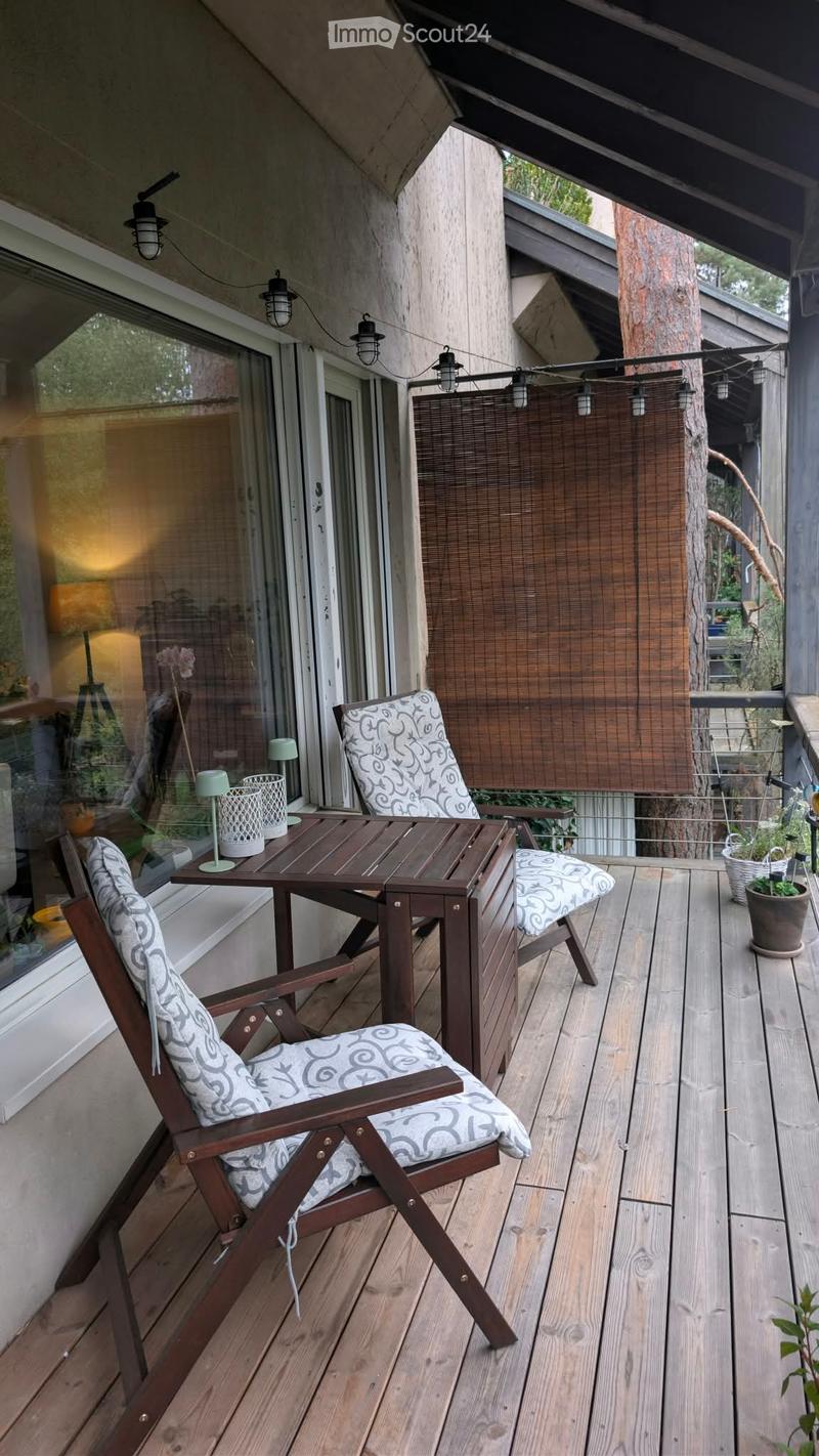 Wooden deck with two wooden chairs with patterned cushions, a small table with a lamp, and potted plants.