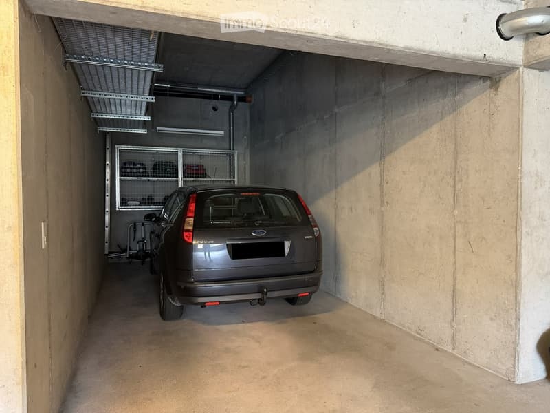 Underground parking space with a single car parked inside, concrete walls and ceiling, storage cage on the left side.