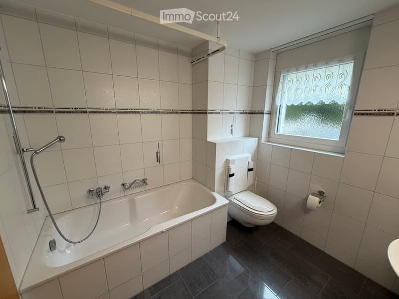 Modern bathroom with a bathtub, toilet, shower hose, and a window with frosted glass. The walls are tiled in light beige with a decorative border, and the floor is dark grey tiles.