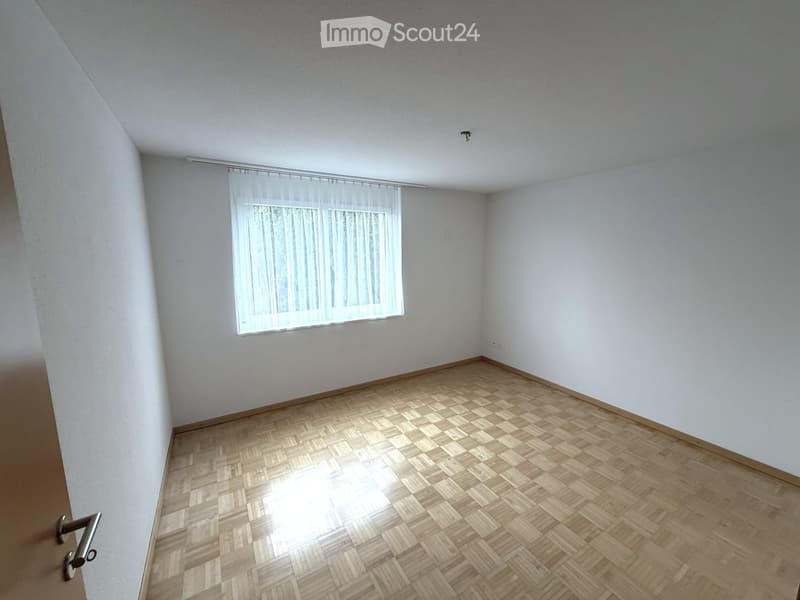 Empty room with wooden parquet flooring, white walls, and a window with sheer curtains.