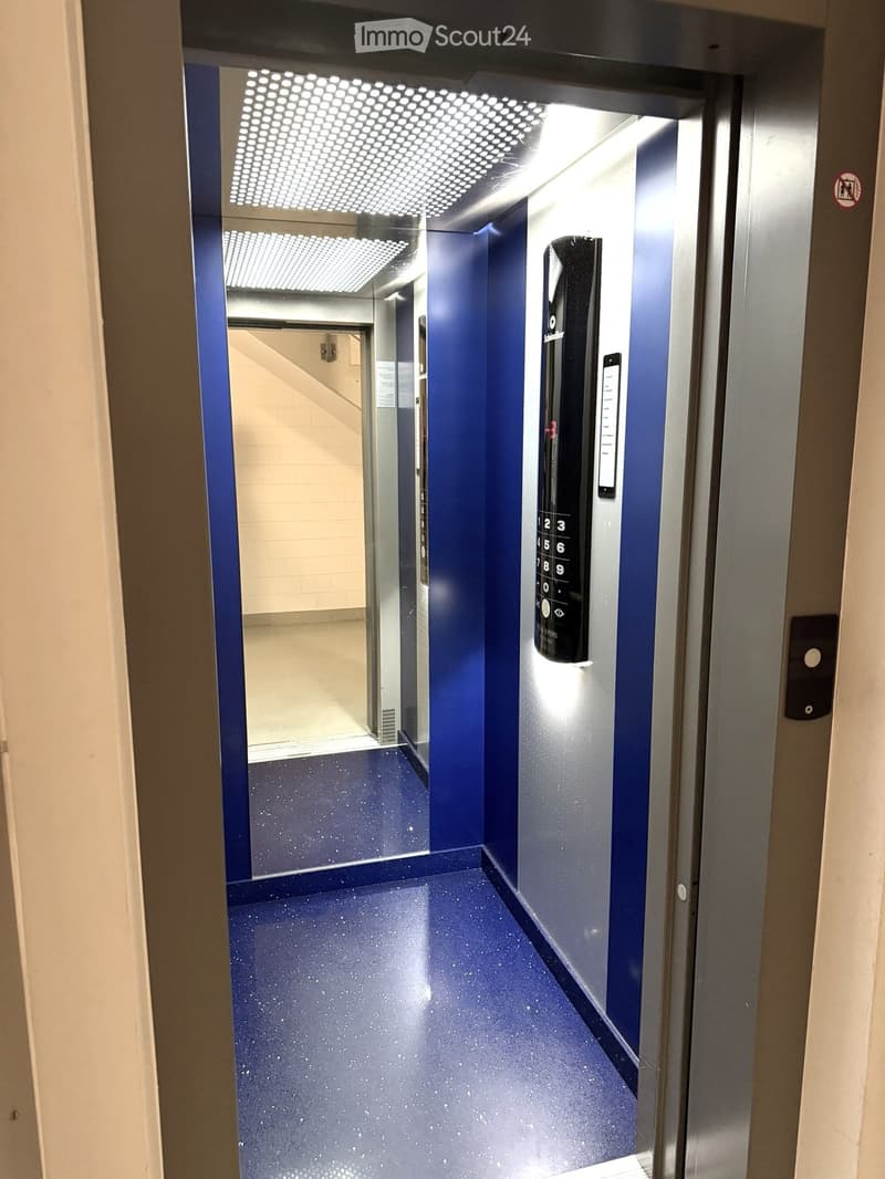 Modern hallway with blue walls and speckled flooring, featuring an elevator control panel and a visible elevator shaft.