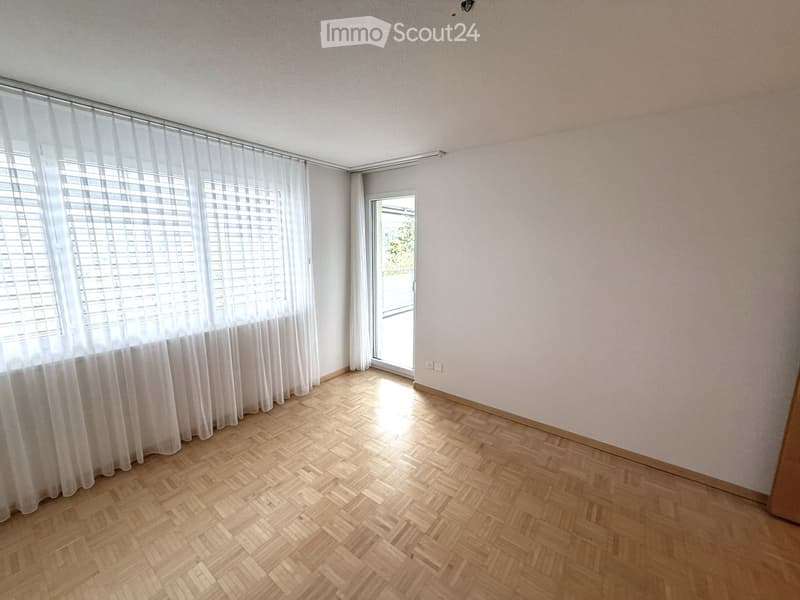 Empty room with wooden herringbone flooring, white walls, large windows with white blinds, and a sliding glass door leading outside.