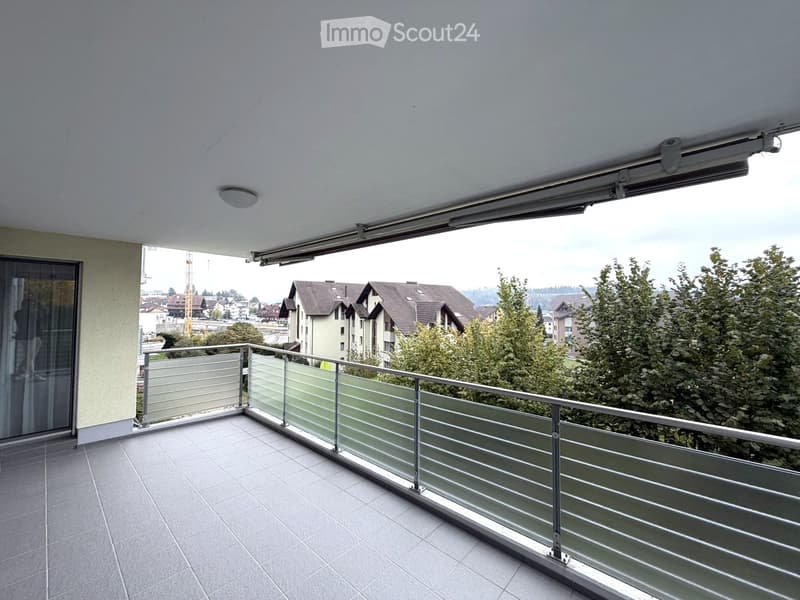 Balcony with modern glass railing, tiled floor, and a retractable awning, overlooking a residential area with trees and buildings.