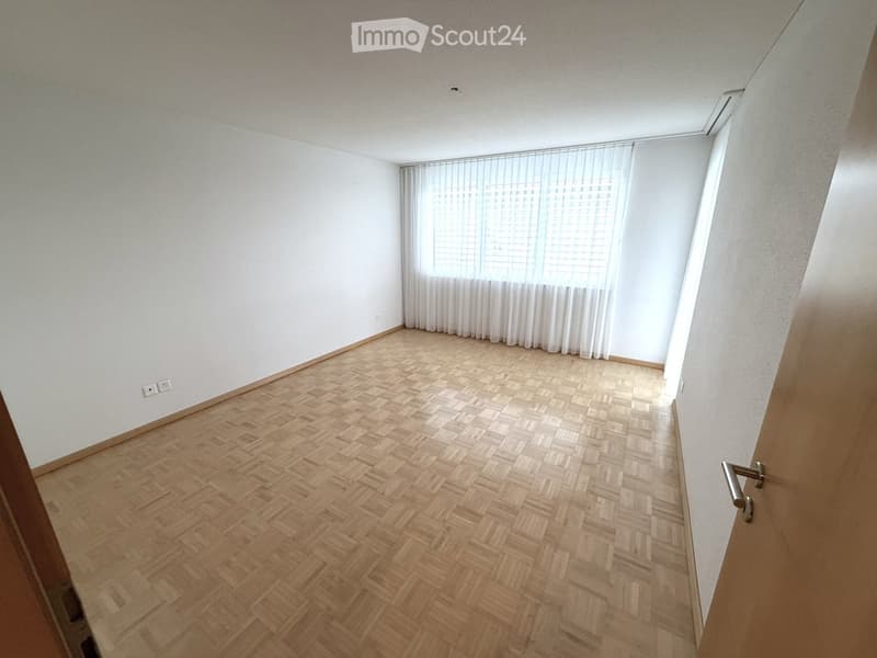 Empty room with wooden herringbone flooring, white walls, and large window with white curtains.