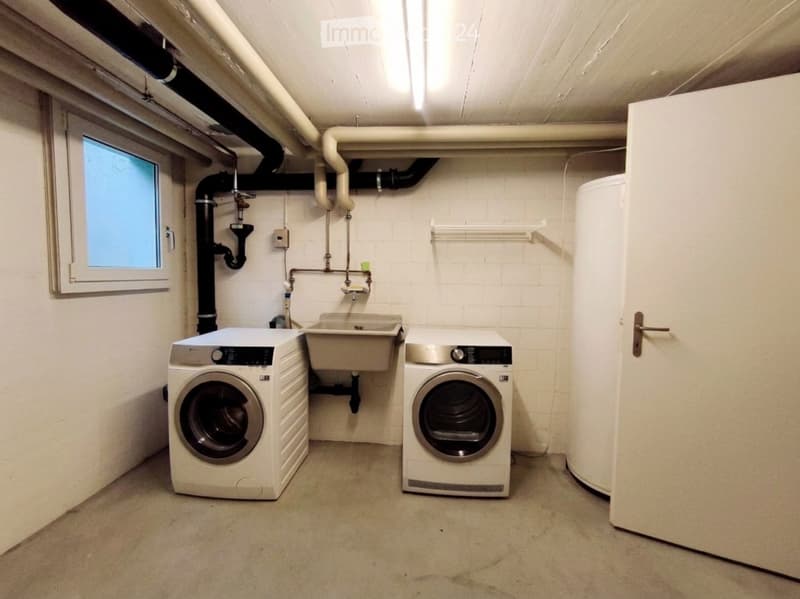 Utility room with a washing machine, dryer, and sink, featuring white walls, exposed pipes, and a small window.