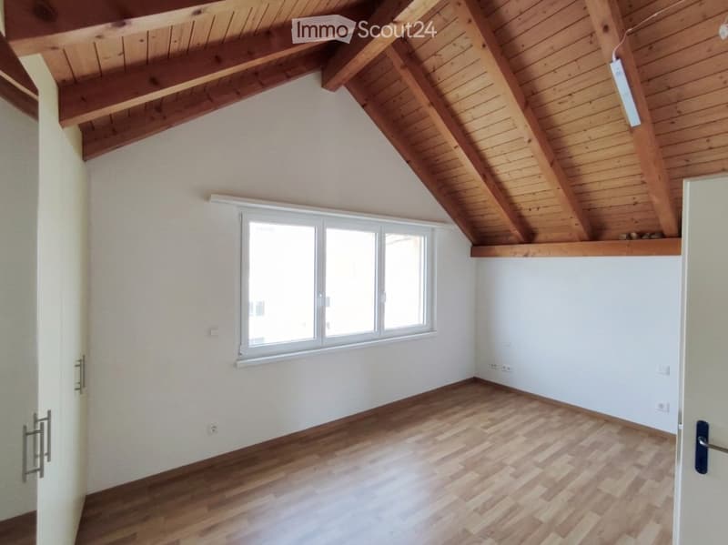 Empty room with wooden flooring, white walls, large window, and exposed wooden ceiling beams.