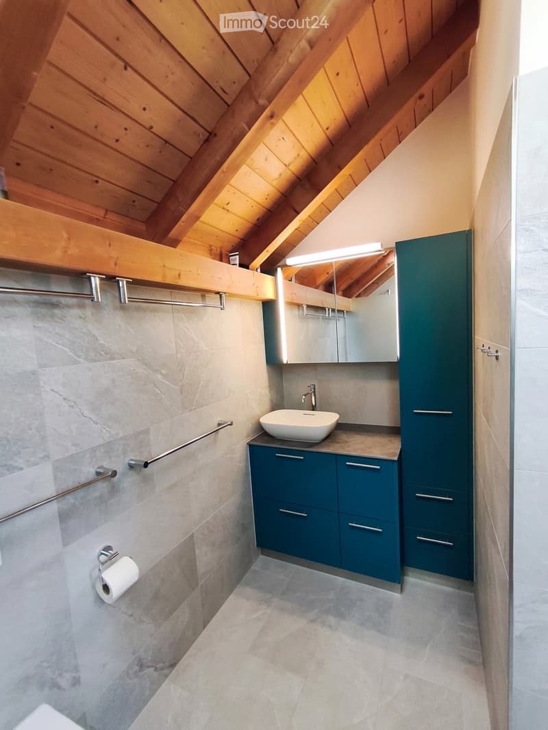 Modern bathroom with wooden slanted ceiling, marble walls, grey tiles, blue cabinets, and a white sink.