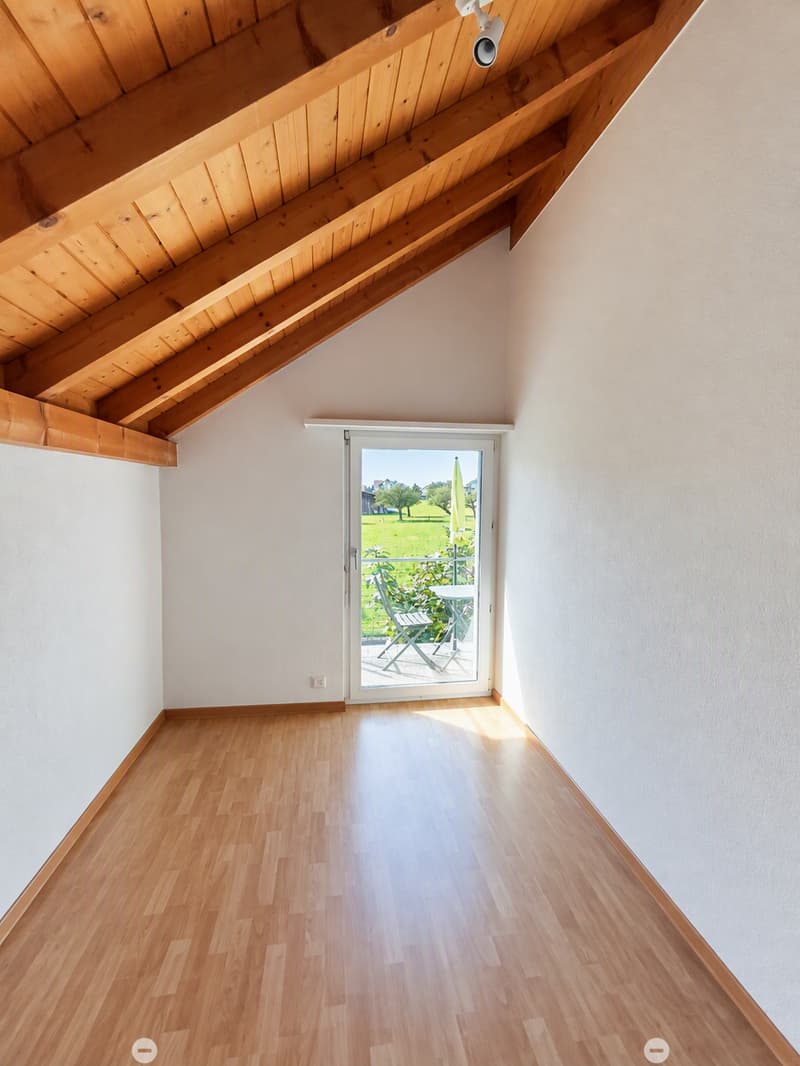 Empty room with wooden flooring, white walls, and a large sliding glass door leading to a balcony or outdoor area. The ceiling has exposed wooden beams.