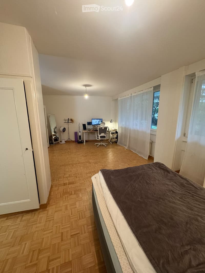 A well-lit bedroom with a wooden floor, a bed with dark bedding, a desk with a computer, a chair, a mirror, and a fan.
