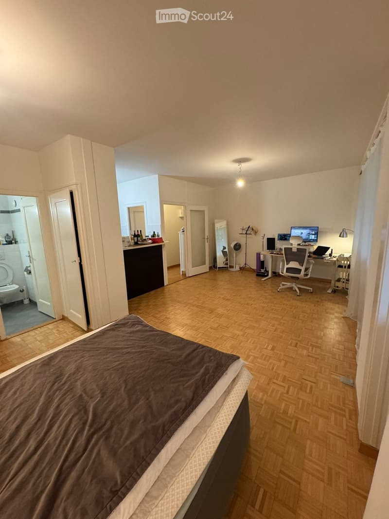 A furnished room with a bed, desk, chair, and a partially visible bathroom. The room features wooden flooring and white walls.