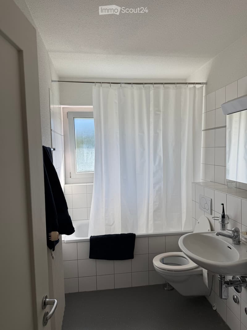 Modern bathroom with white tiles, a bathtub, shower curtain, toilet, sink, and a towel hanging on the wall.
