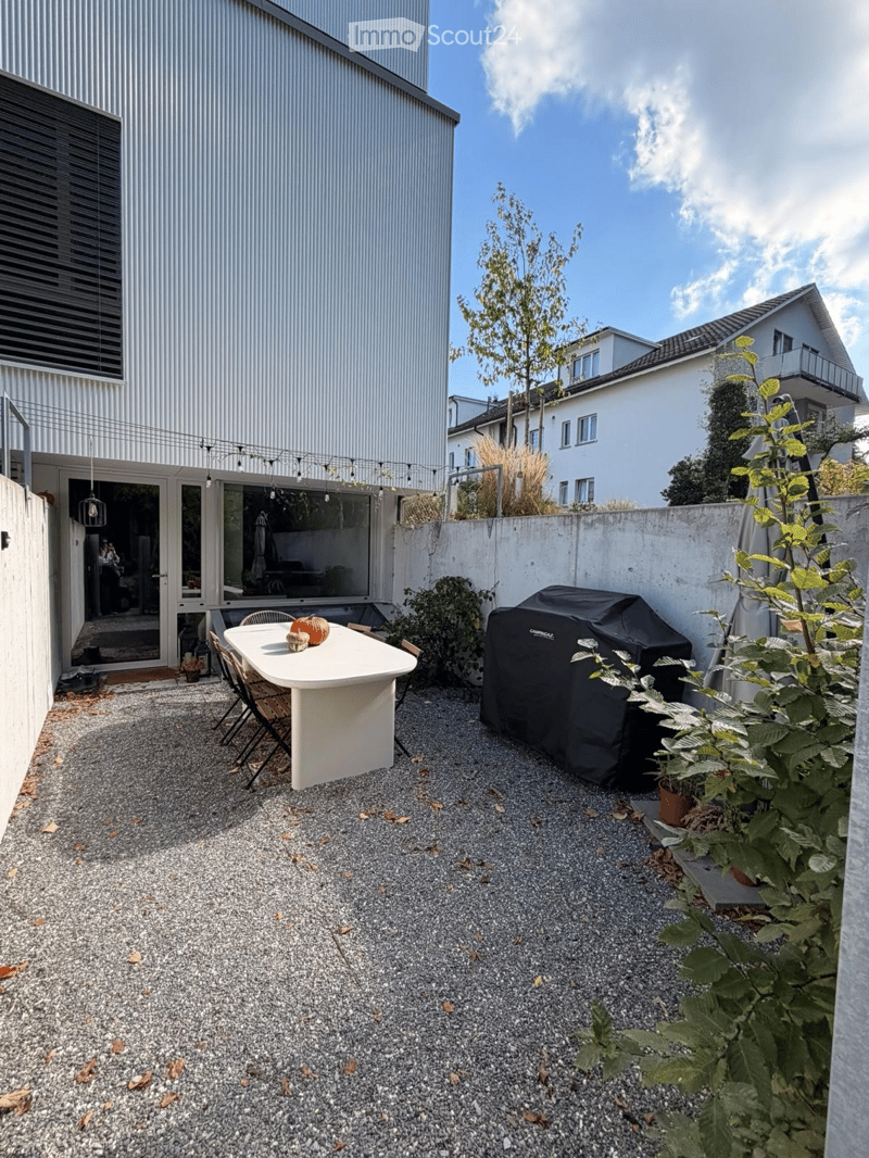 Compact outdoor patio with a white table, chairs, a covered grill, and potted plants, adjacent to a modern building.