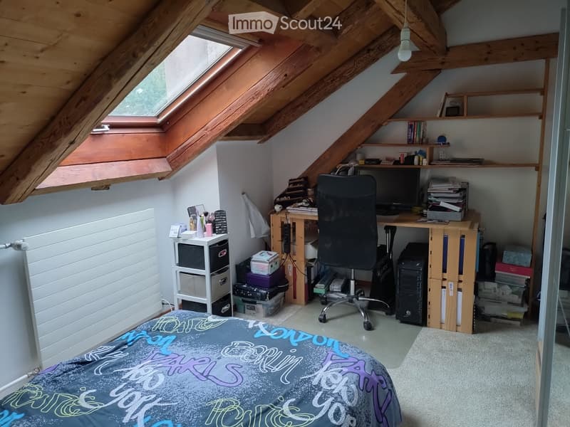 Cozy attic bedroom with slanted wooden ceilings, skylight windows, a bed with a colorful duvet, a desk with a chair, shelves, and storage boxes.