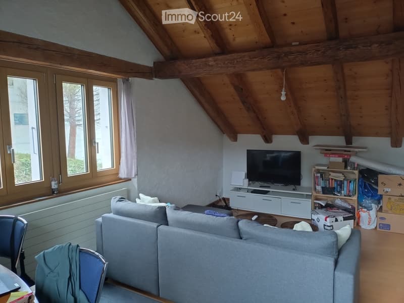 Living room with exposed wooden beams, large windows, grey sofa, coffee table, TV stand, bookshelf, and a laptop on a table.