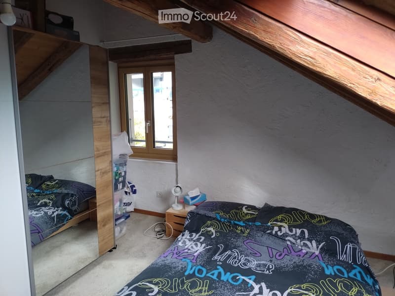 Cozy attic bedroom with slanted wooden ceilings, two single beds with colorful bedding, a nightstand with a lamp, and a shelf with storage boxes.