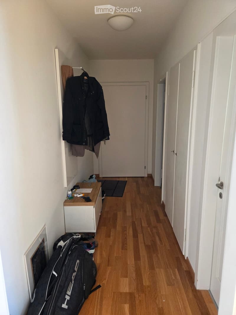 A narrow hallway with wooden flooring, white walls, and several doors. A small wooden bench with various items, including a jacket, a backpack, and some small objects, is placed against the wall. A coat rack with a jacket and a towel is also visible.