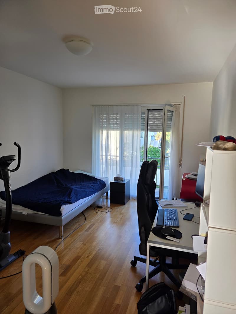 A well-lit bedroom with a bed, desk, chair, and exercise bike. The room features wooden flooring, a large window with white curtains, and a nightstand with a lamp.