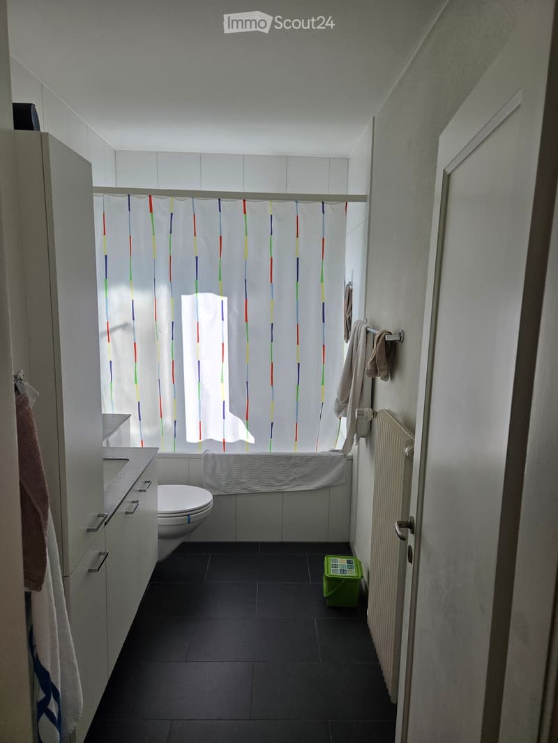 Modern bathroom with a toilet, bathtub, shower curtain, towels, radiator, and a small green stool on the floor.