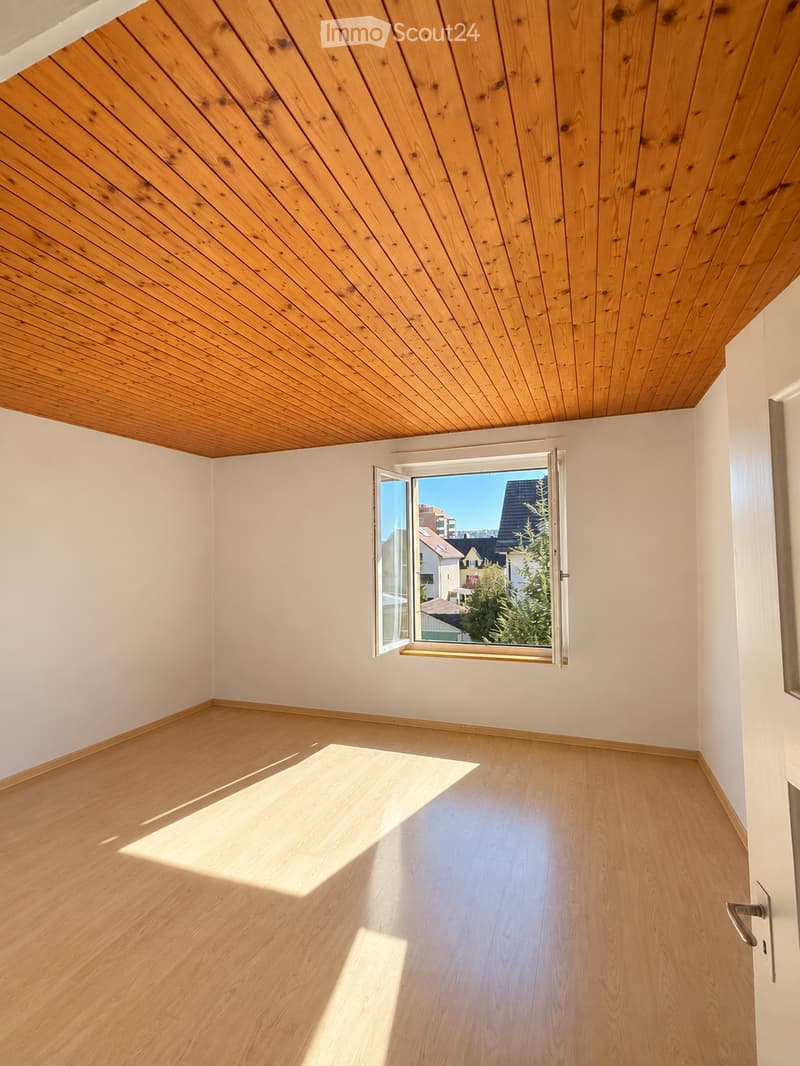 Empty room with light wooden flooring and white walls, featuring a large window with a view of the outside and a wooden slanted ceiling.