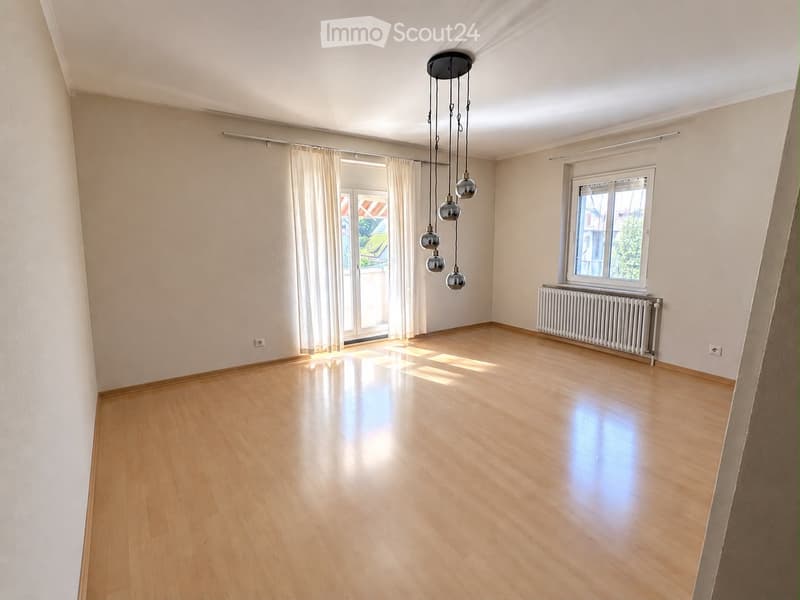 Empty room with wooden flooring, white walls, large windows with curtains, and a hanging light fixture.