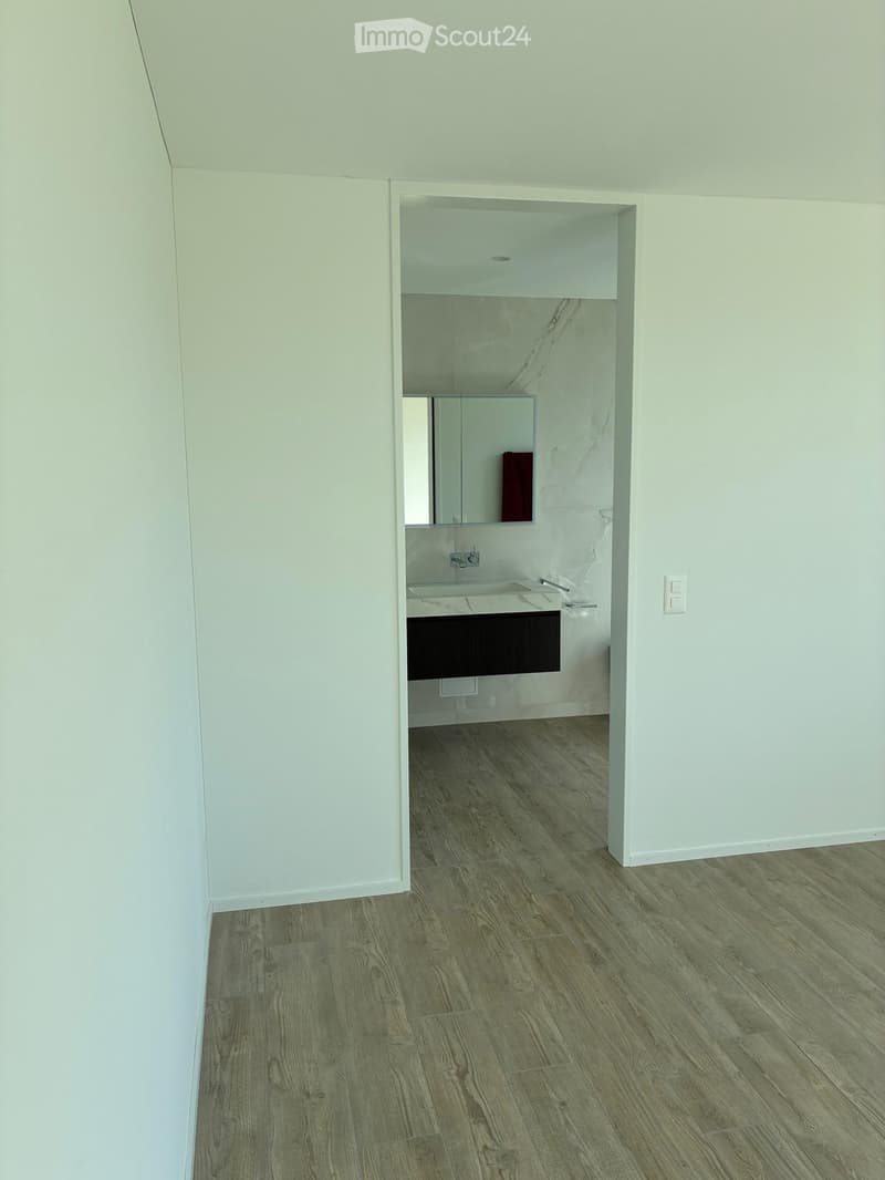 Empty hallway leading to a small, modern bathroom with white walls, marble tiles, and a wooden floor. The bathroom features a sink, mirror, and dark cabinetry.