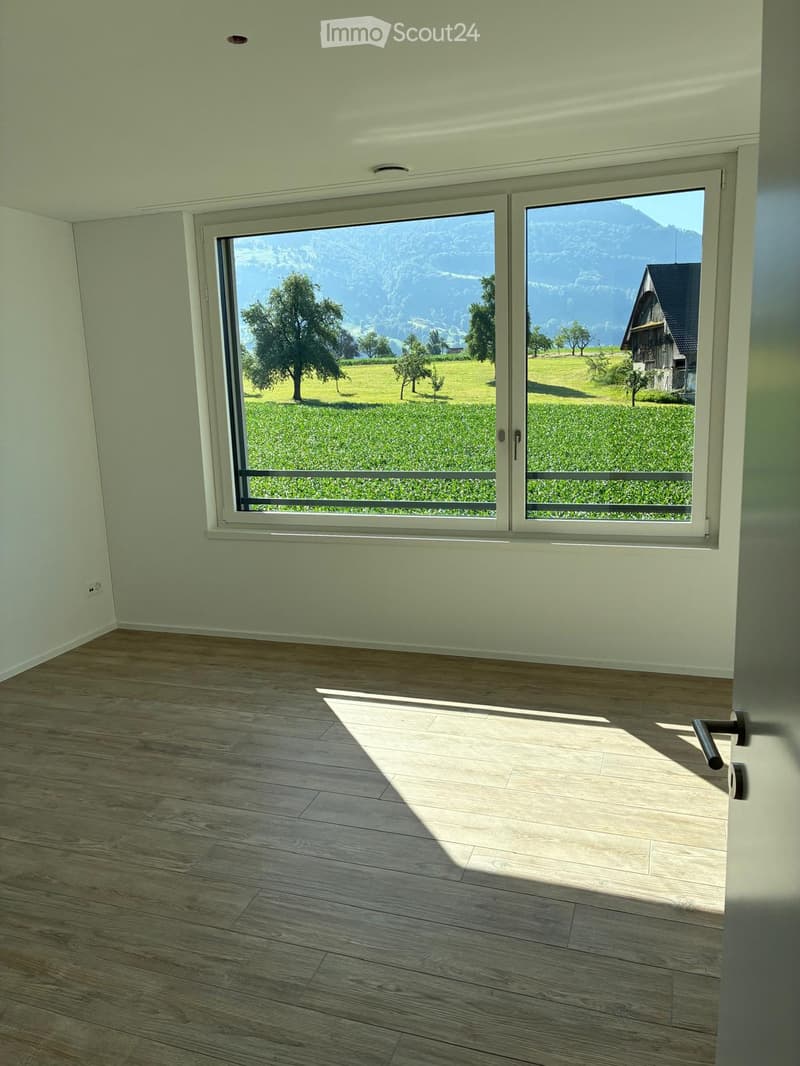Empty room with light-colored wooden flooring and large windows offering a scenic view of a green field and mountains.