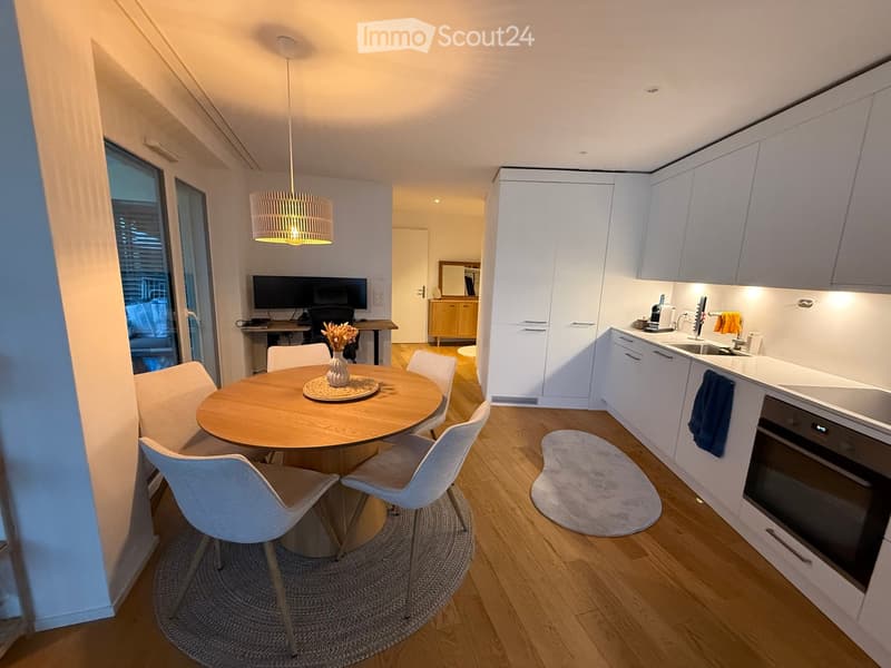 Modern kitchen and dining area with white cabinets, a round wooden dining table with four white chairs, a sink, oven, and a small workspace with a screen.