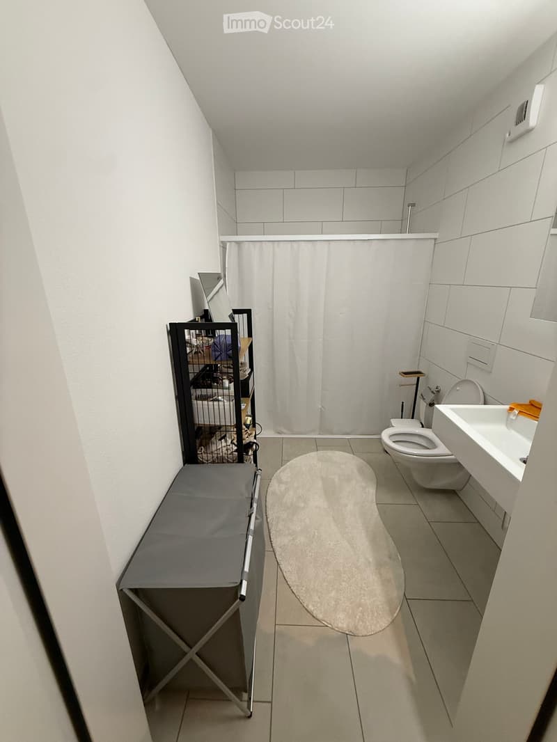 Modern bathroom with white tiles, a toilet, sink, mirror, shower curtain, bath mat, and a small cage with a table nearby.