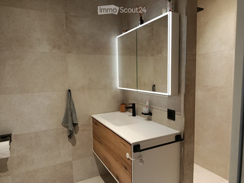 Modern bathroom with a white countertop sink, wooden cabinet, illuminated mirror, and a towel hanging on the wall.