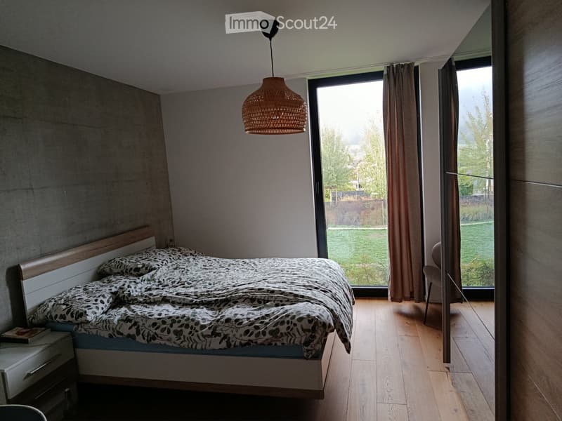 Modern bedroom with wooden flooring, large window with view, bed with patterned bedding, nightstand, chair, and pendant light.