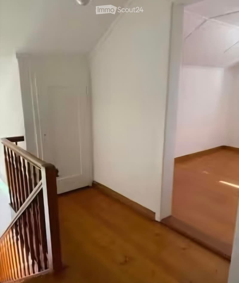 Empty hallway with wooden flooring, white walls, and a staircase with a wooden railing on the left side.