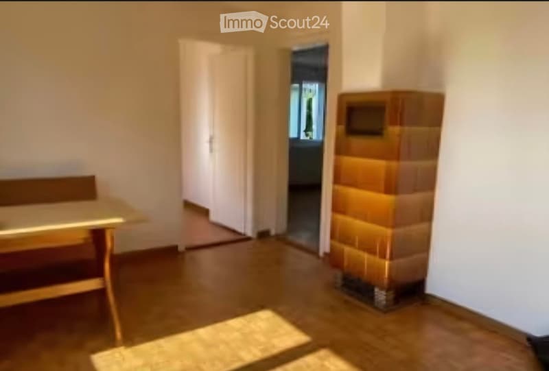 A small, furnished room with a wooden table, a chair, and a bench. The room has wooden flooring, white walls, and a fireplace with a decorative brick surround. A doorway leads to another room with a visible window.