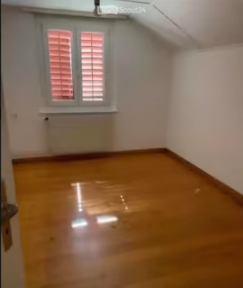 Empty room with wooden flooring and white walls, featuring a window with blinds and a door on the left side.