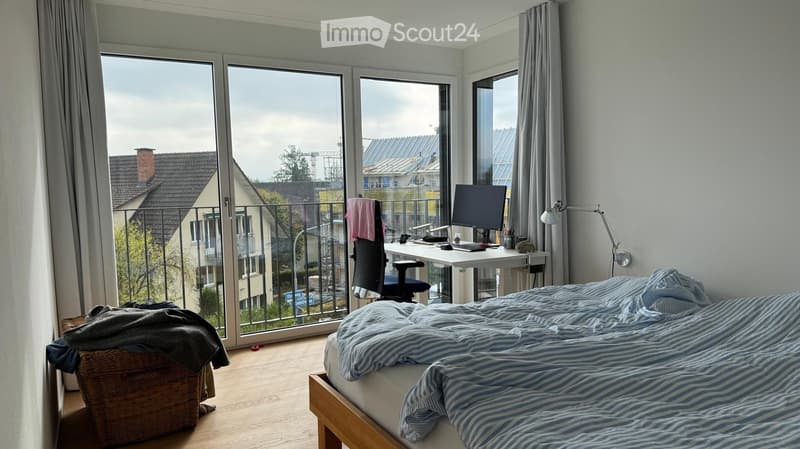 Modern bedroom with a large window and balcony access, featuring a bed with striped bedding, a desk with a computer, and a wicker basket on the floor.