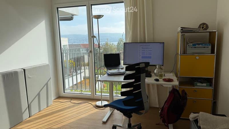 Bright room with a desk, chair, laptop, screen, and a view of the city and water through large windows.