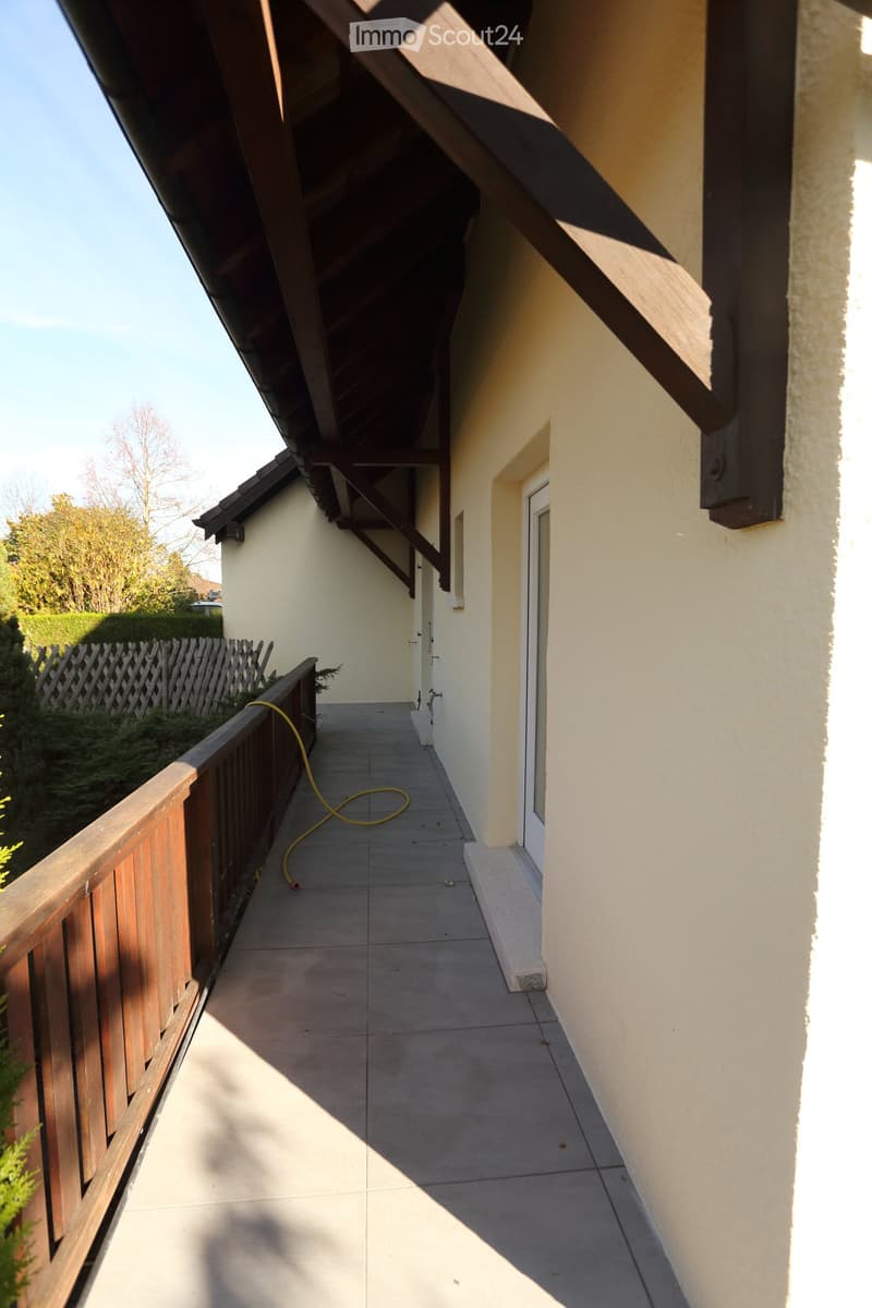 A narrow outdoor corridor with tiled flooring, wooden railing on one side, and a covered roof supported by wooden beams. The corridor leads to a door and is bordered by a wooden fence and greenery.