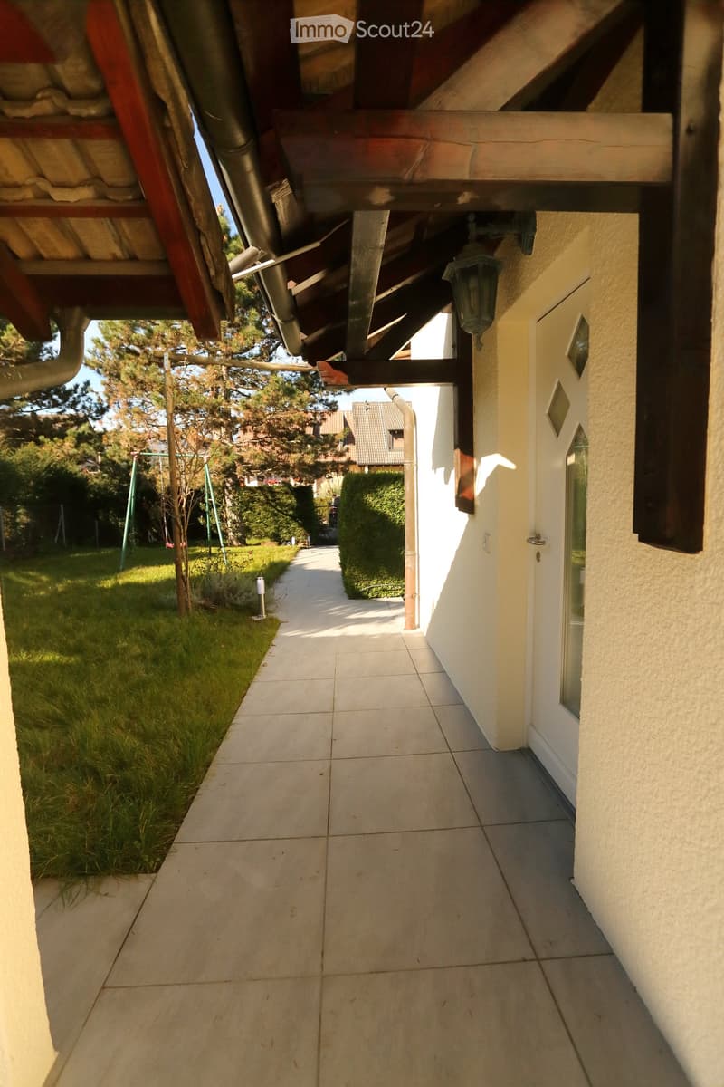 A covered pathway leading to a house entrance, with tiled flooring, a grassy area to the left, and a swing set visible in the background.