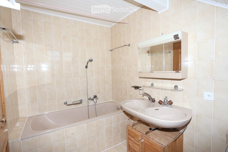 Bathroom with beige tiled walls and floor, featuring a bathtub with a handheld shower, a double sink on a wooden cabinet, a large mirror with built-in lighting, and a handrail for safety.