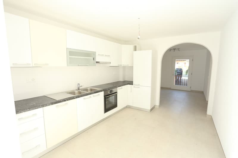 Modern, unfurnished kitchen with white cabinets, a stainless steel sink, and an oven. The space features a dark countertop and light tiled flooring, with an arched doorway leading to another room.
