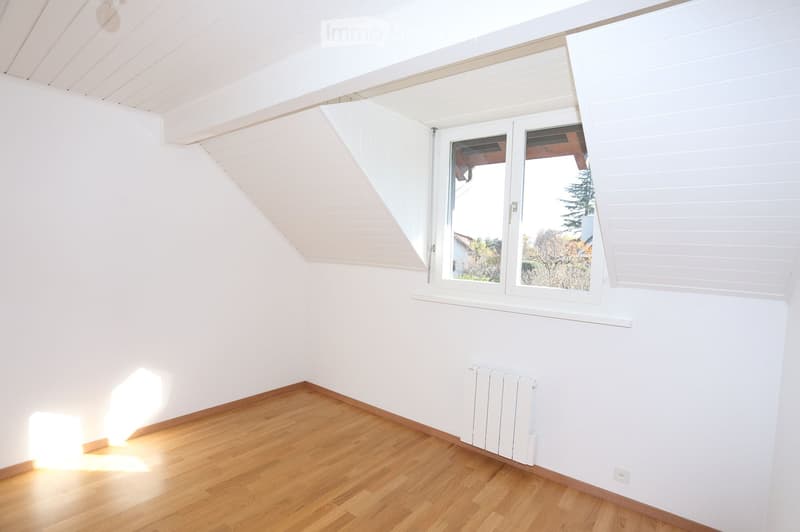Empty room with wooden flooring, white walls, and a large window with white frames. The room has a slanted ceiling and a radiator on the wall.
