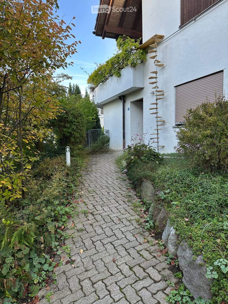 A pathway leads to a modern white house with a wooden roof and a small balcony. The garden is lush with various plants and trees, and the house features a decorative wooden structure on the wall.