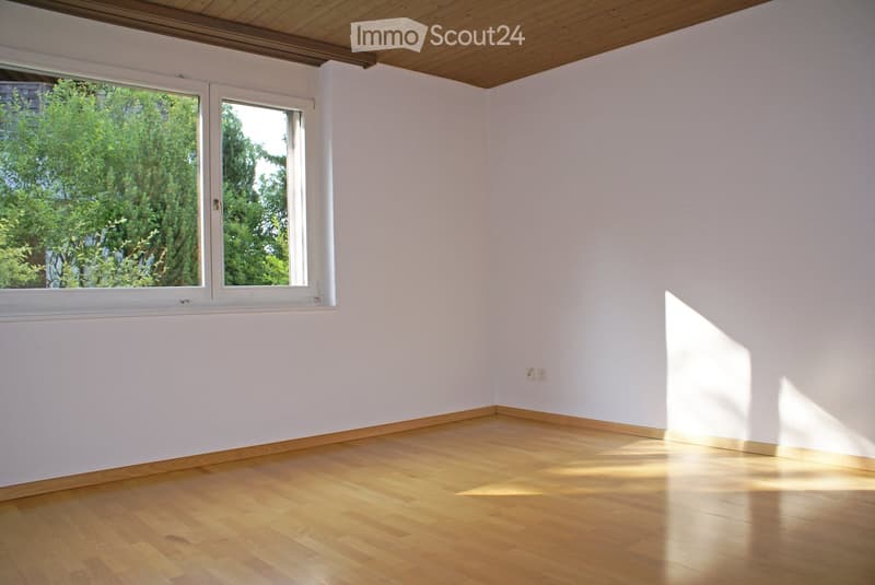 Empty room with light wooden flooring, white walls, and large windows allowing natural light to enter. The ceiling is wooden, and there are visible electrical outlets on the wall.