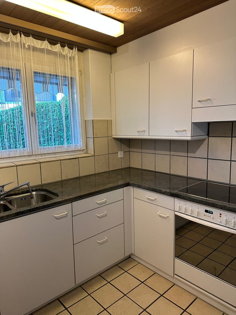 Modern kitchen with white cabinets, black countertop, stainless steel sink, and built-in oven. Features tiled backsplash and window with sheer curtains.