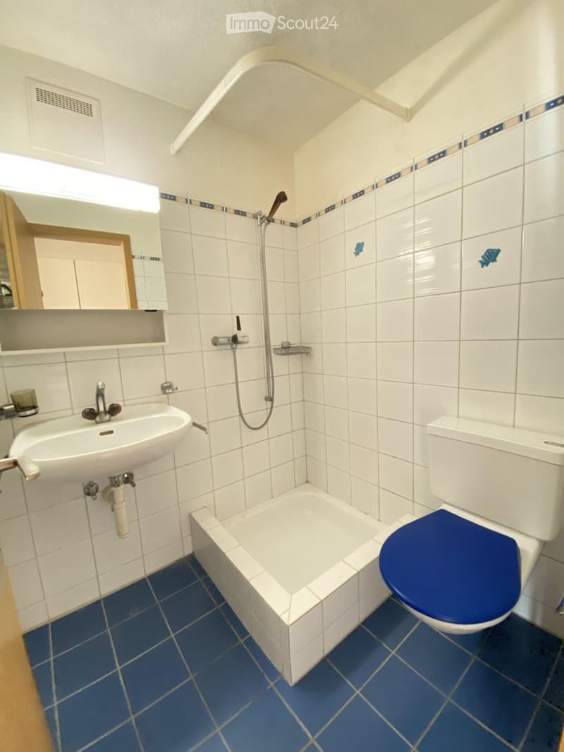 Bathroom with white tiled walls, blue tiled floor, white sink, toilet with blue seat, bathtub, and a mirror above the sink.